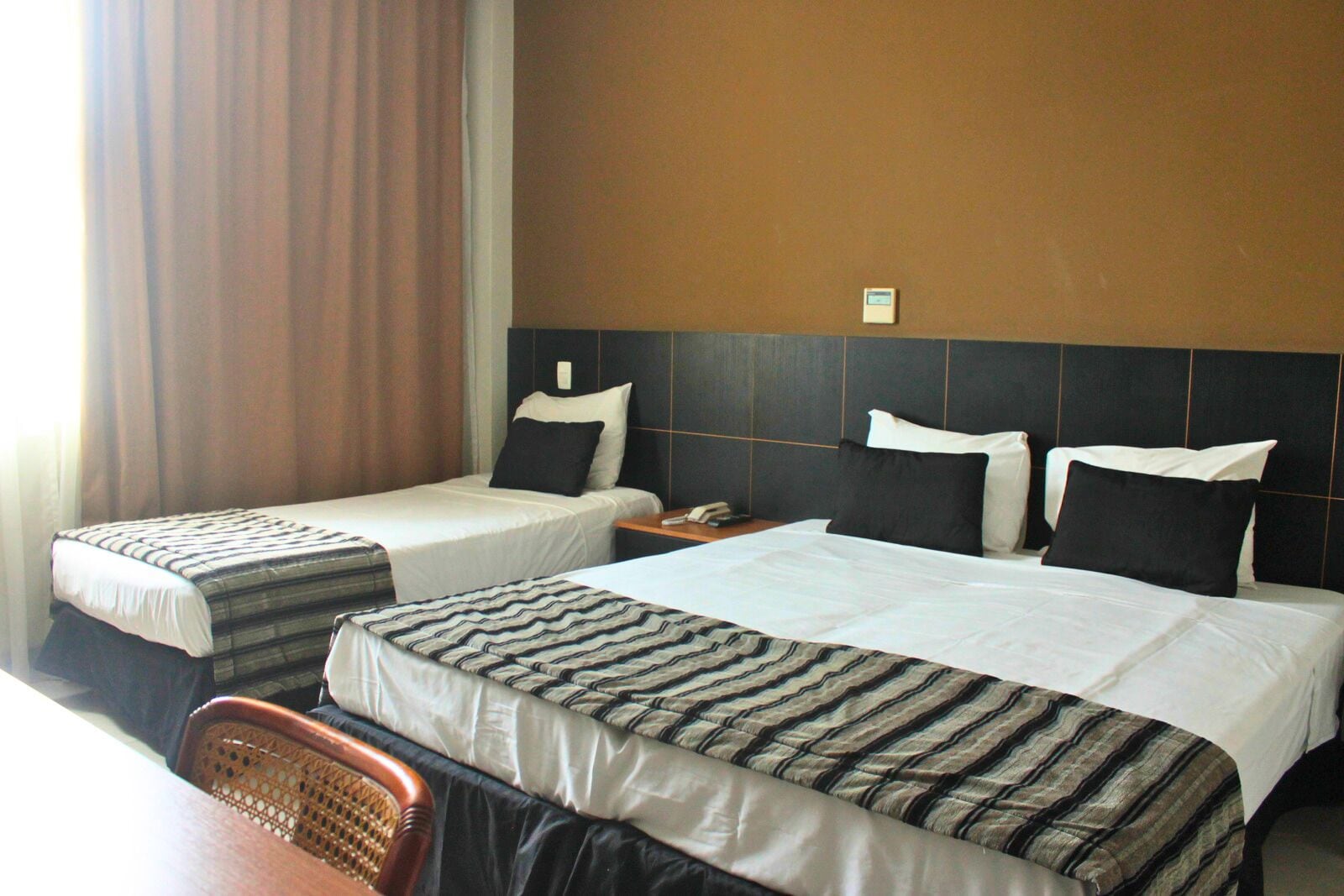 A hotel room with two single beds and a double bed, each with striped bedspreads. There is a wooden chair and a nightstand with a phone.