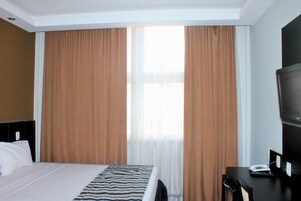 Habitación de hotel con cama, escritorio, computadora y teléfono, además de cortinas grandes.