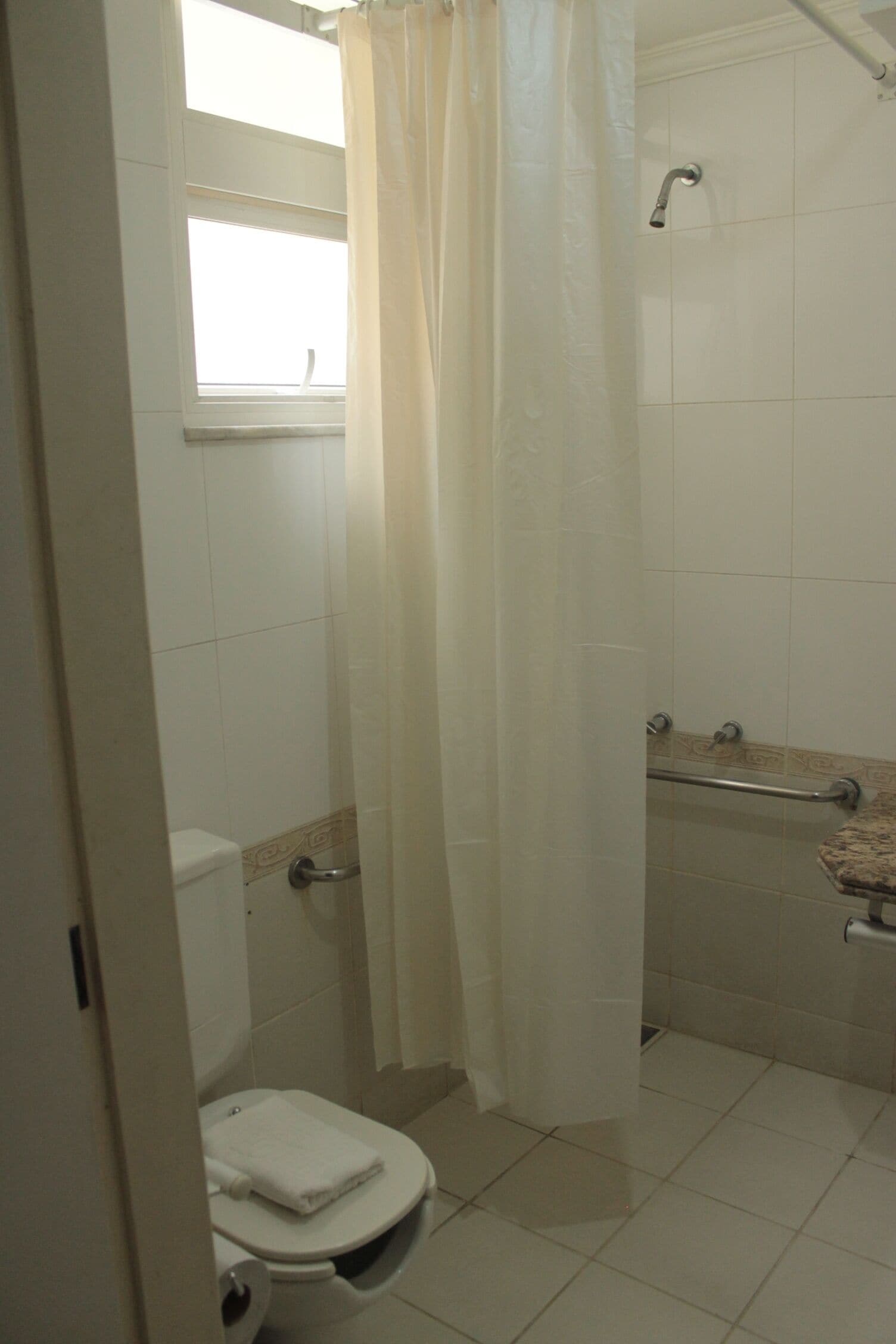 A bathroom with a toilet, a shower with a grab bar, and a window with a curtain.
