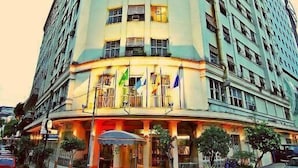 A tall, multi-story hotel with a curved corner facade, balconies, and a well-lit entrance area with potted plants. - Hotel Atlântico Tower - SOFT OPENING (Rio de Janeiro)