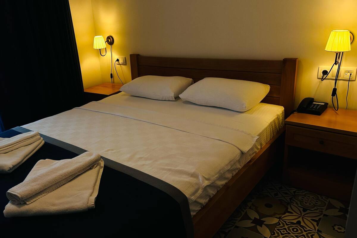 A hotel room with a bed, two bedside lamps, a nightstand with a phone, and a window with curtains.