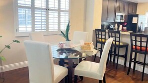 Un comedor con una mesa de vidrio rodeada de sillas blancas, una araña y vistas a una cocina con armarios oscuros y un microondas.