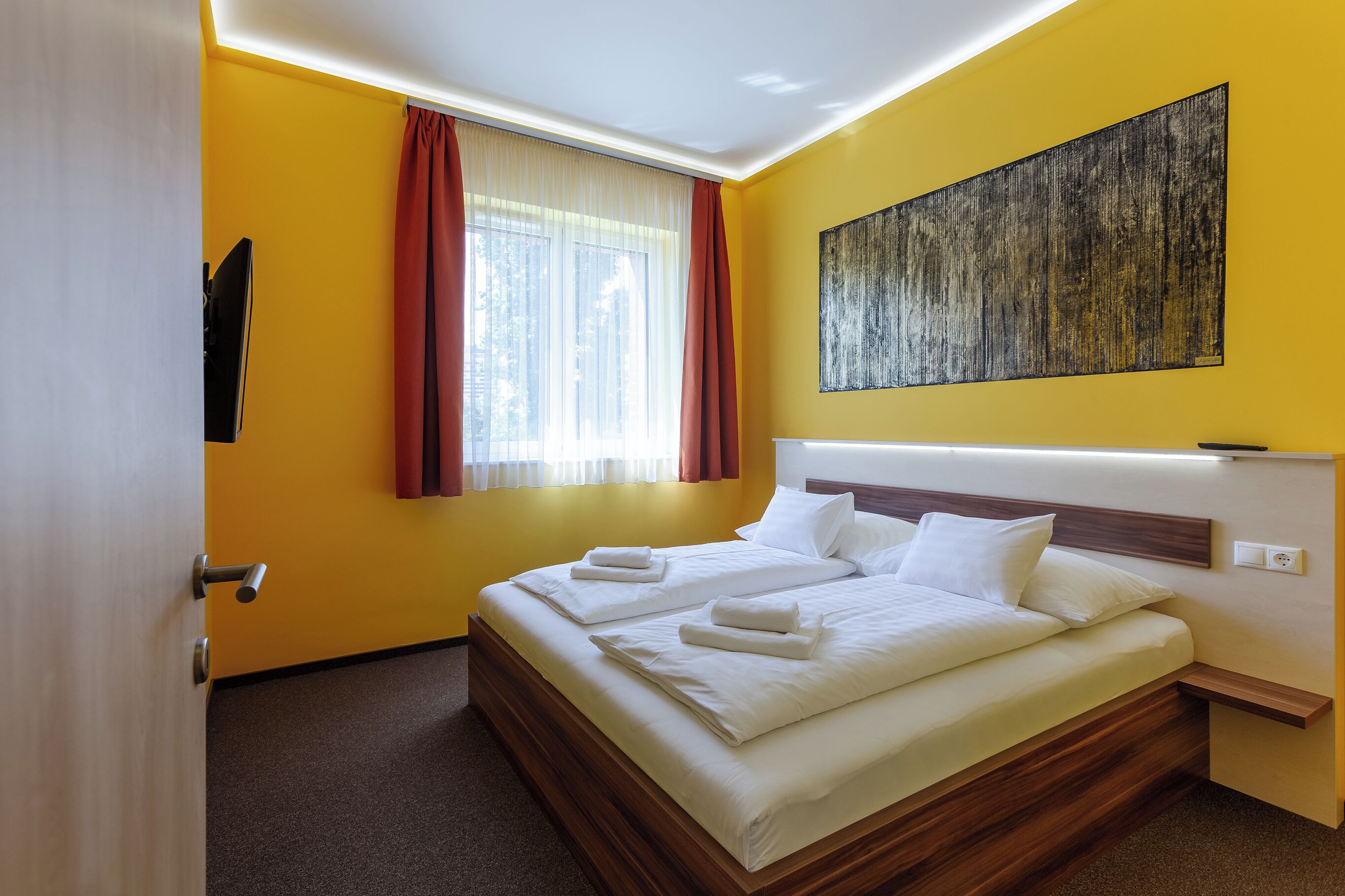 Quarto de hotel com uma cama grande, cabeceira, janela com cortinas e um quadro na parede.