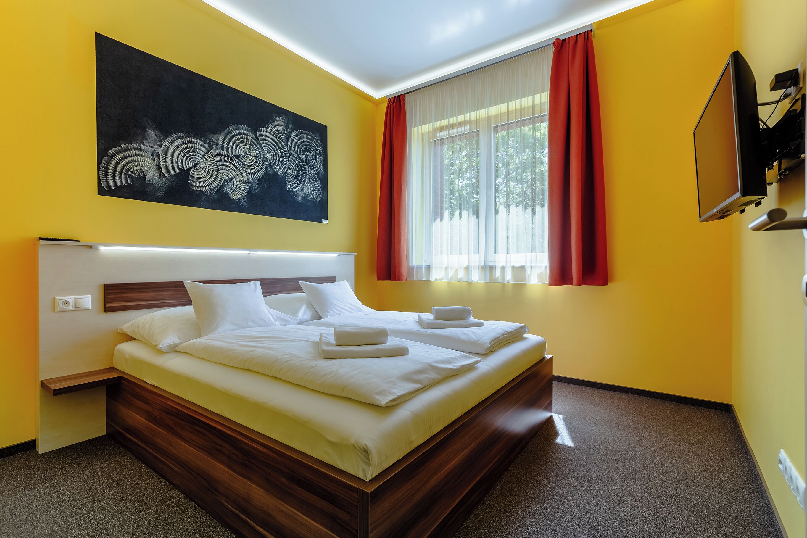 Una cama doble con sábanas blancas, una mesita de noche, un televisor montado en la pared y una ventana con cortinas rojas.