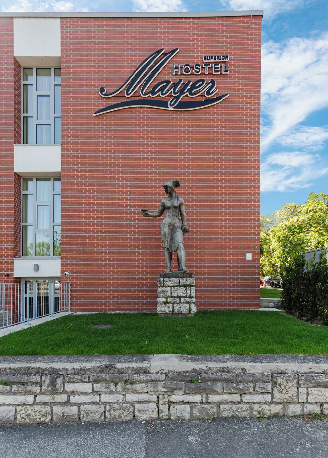 A brick building with a sign reading "Superior Hostel Mayer" and a statue of a woman in front.