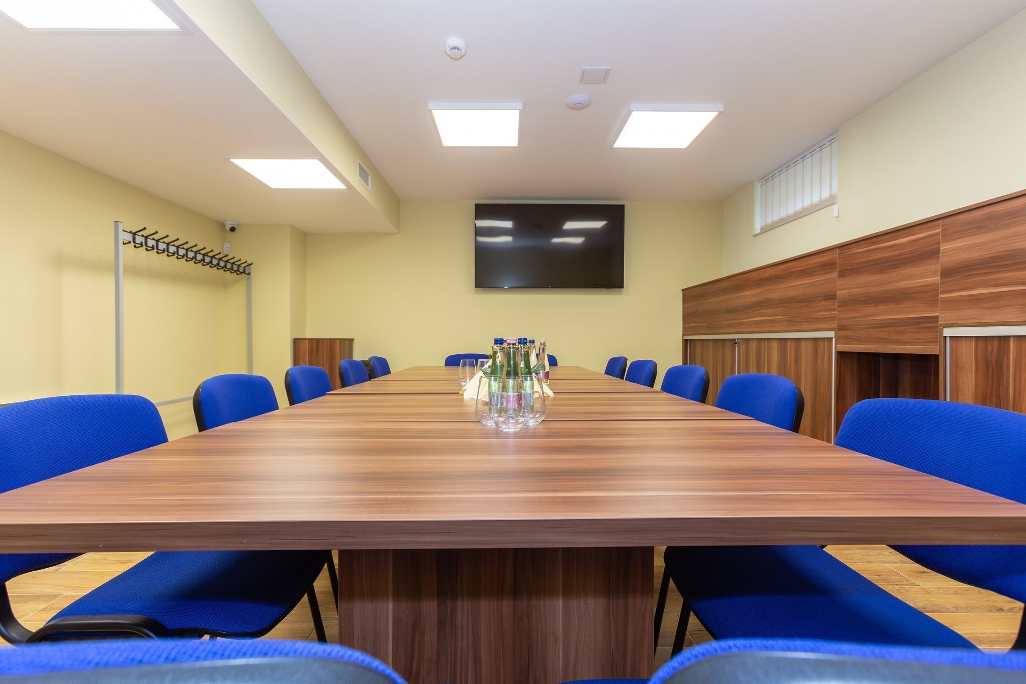 Un salón de conferencias con una larga mesa de madera, sillas azules, un televisor de pantalla plana y una cámara de seguridad en el techo.