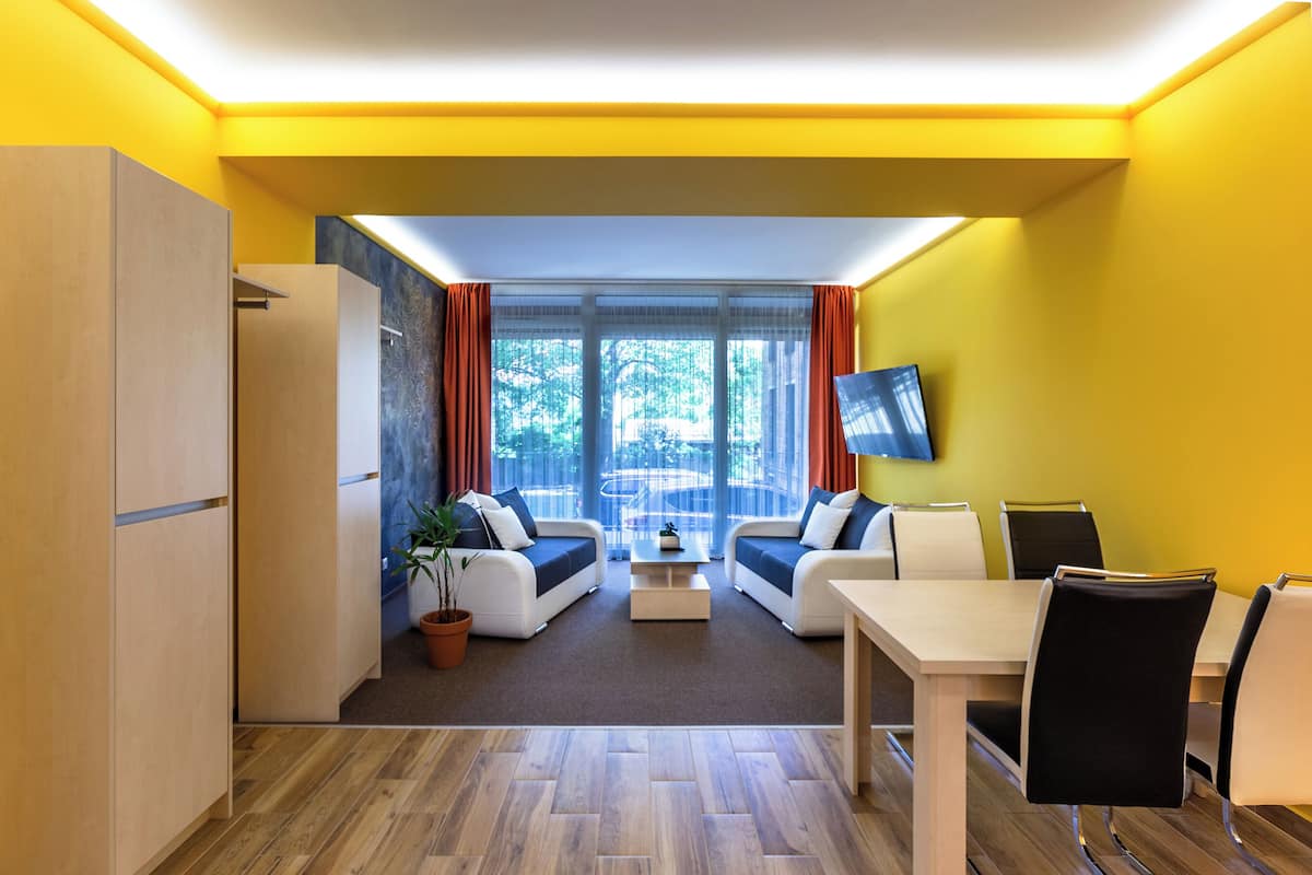A modern living room with a sofa set, a dining table, and chairs. There is a television mounted on the wall and a window with curtains.
