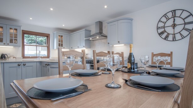A modern kitchen with light blue cabinets, a wooden dining table set with plates and wine glasses, and a large wall clock.