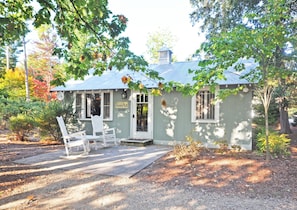Charming cottage with a porch and rocking chairs, surrounded by lush trees and shrubs.