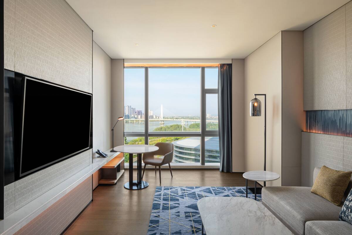 Une chambre d’hôtel moderne dotée d’une grande télévision, d’un canapé, d’une petite table et offrant une vue sur un pont et le paysage urbain.