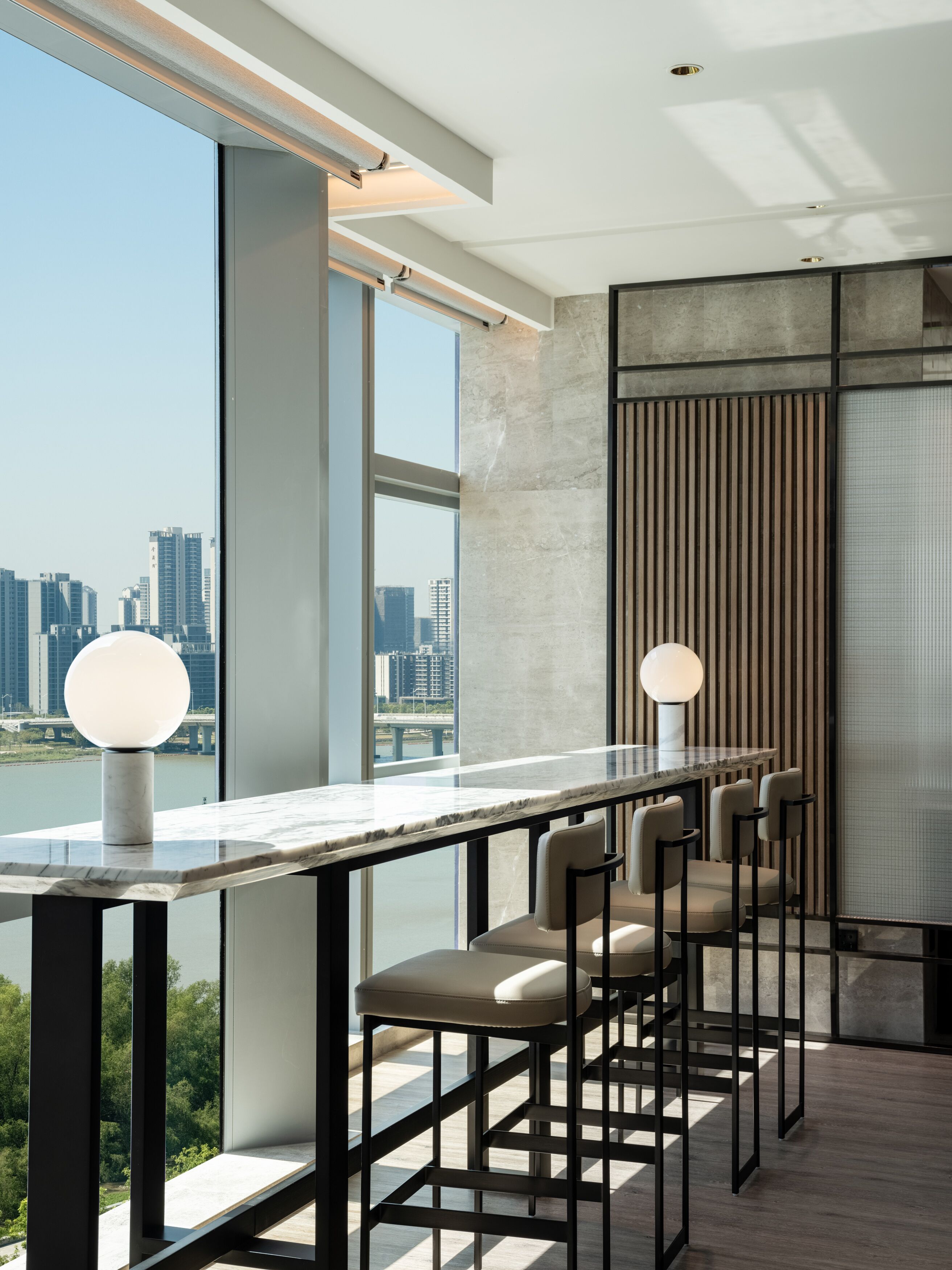 A modern bar area with a long counter, multiple bar stools, and large windows offering a view of a cityscape and river.