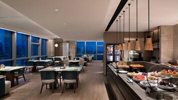 A modern dining area with tables and chairs, a buffet with various food items, and large windows offering a city view.