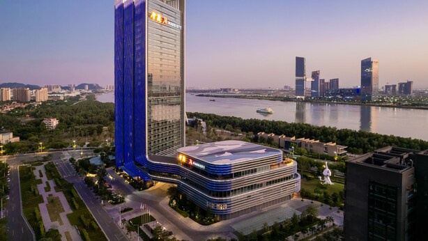 Un hotel moderno con una imponente torre vertical y un edificio más bajo con techo curvo, situado cerca de una masa de agua con un paisaje urbano de fondo.