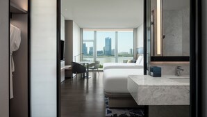 Una habitación de hotel moderna con una cama grande, una zona de estar con escritorio y silla, un baño con espejo grande y lavamanos, y vistas a la silueta urbana a través de amplias ventanas.