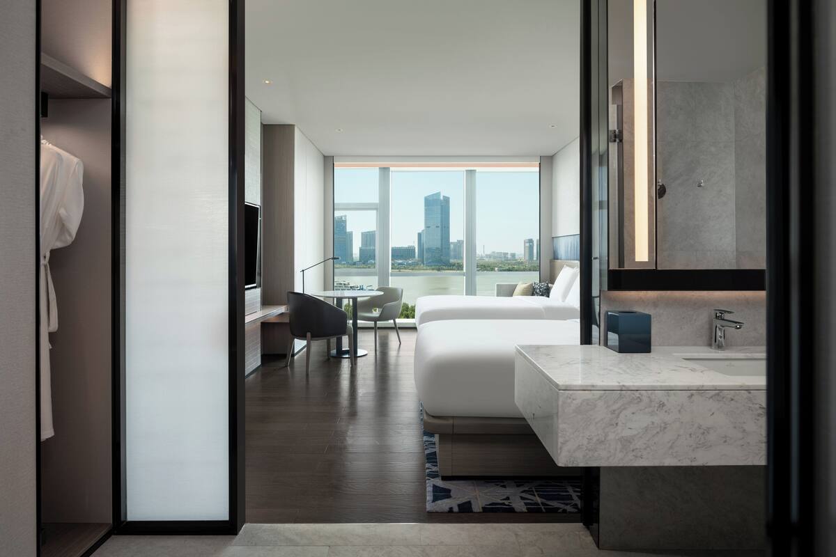 Une chambre d’hôtel moderne dotée d’un grand lit, d’un coin salon avec un bureau et une chaise, d’une salle de bain avec un grand miroir et un lavabo, et offrant une vue sur le panorama urbain grâce à de grandes fenêtres.