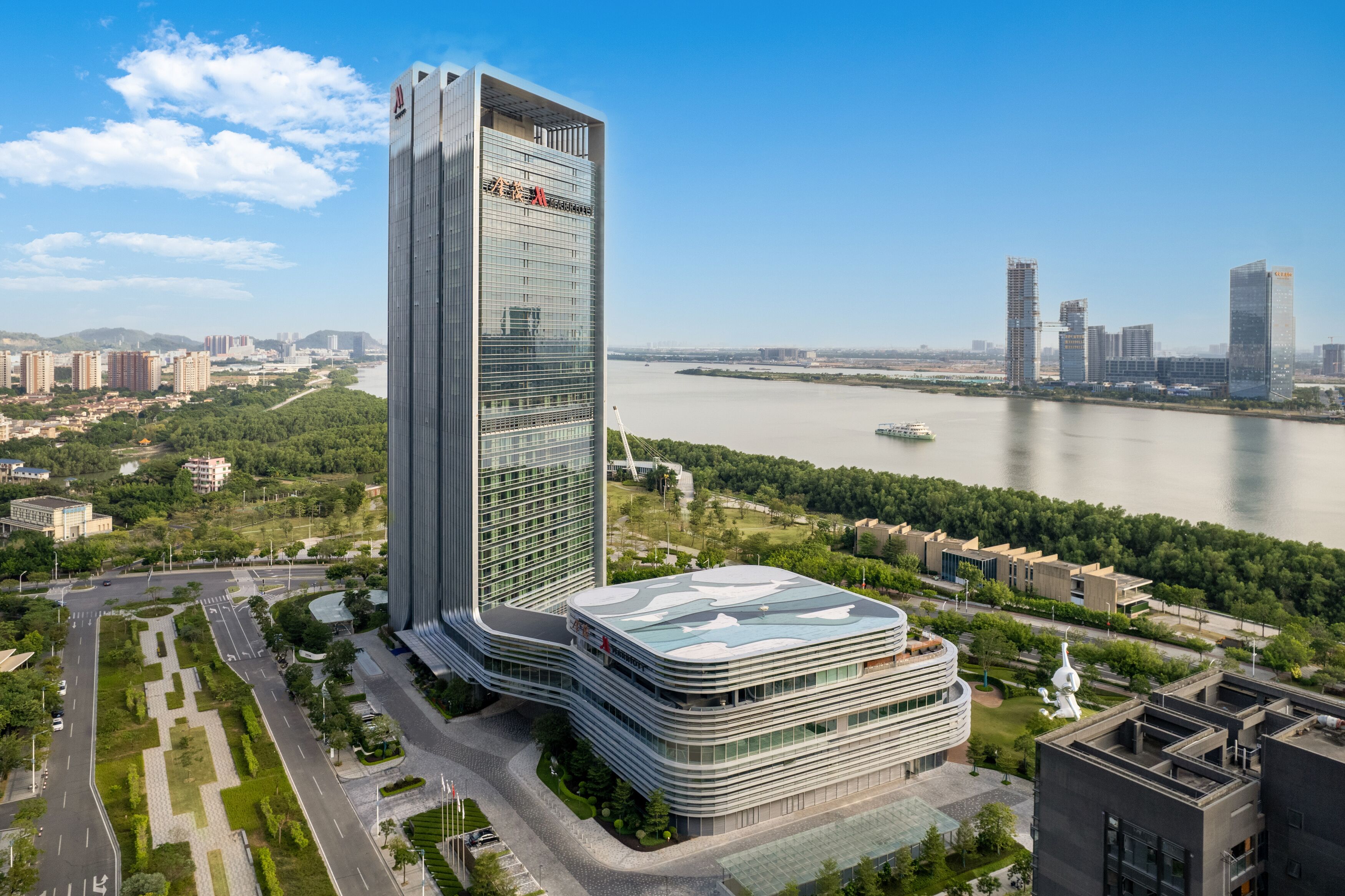 A modern high-rise building with a glass facade, surrounded by greenery and a river.