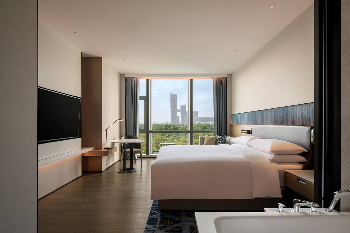 Une chambre d’hôtel moderne avec un grand lit, un téléviseur fixé au mur, une petite table et une vue sur les toits de la ville.