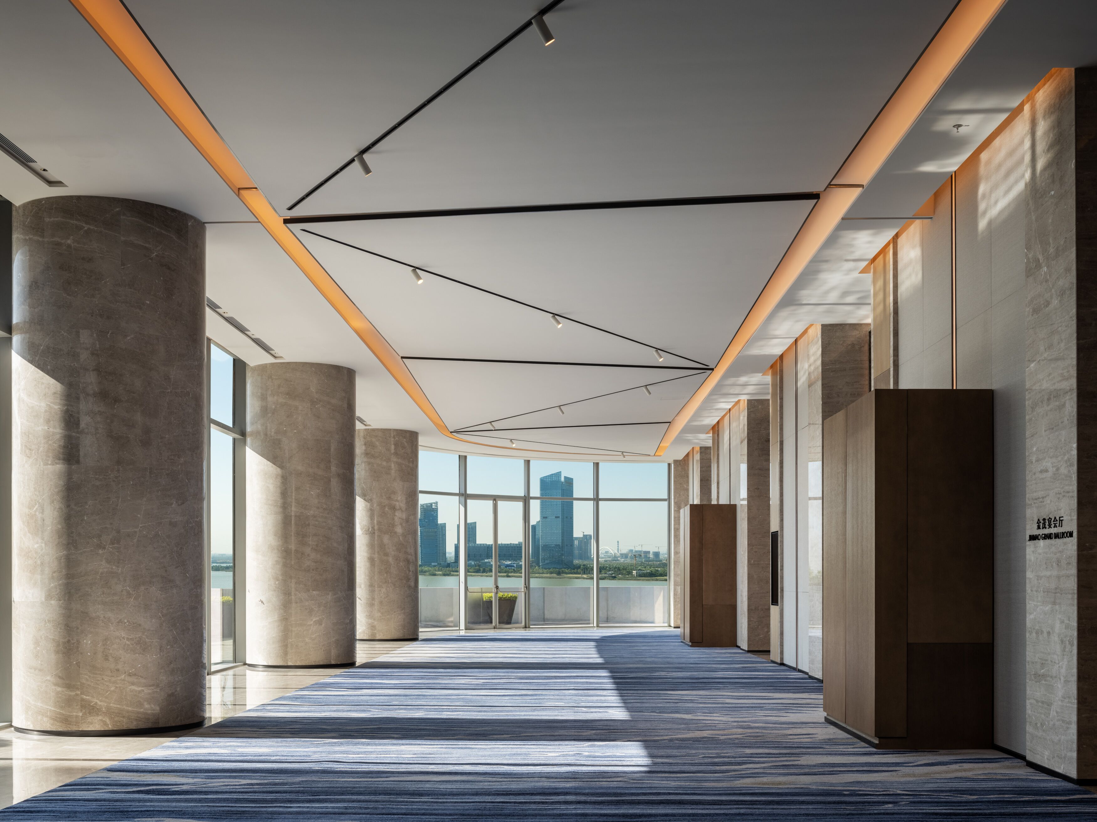 A modern hotel corridor with large windows, concrete pillars, and a patterned carpet.