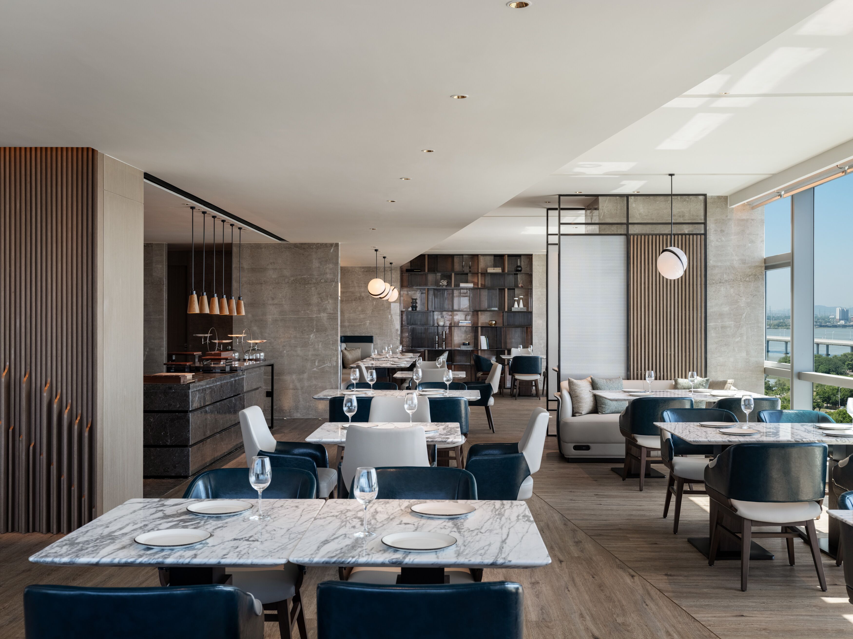 Ein modernes Restaurant mit Marmortischen und blauen Stühlen, mit einer Küchenzeile und Blick auf eine Brücke.