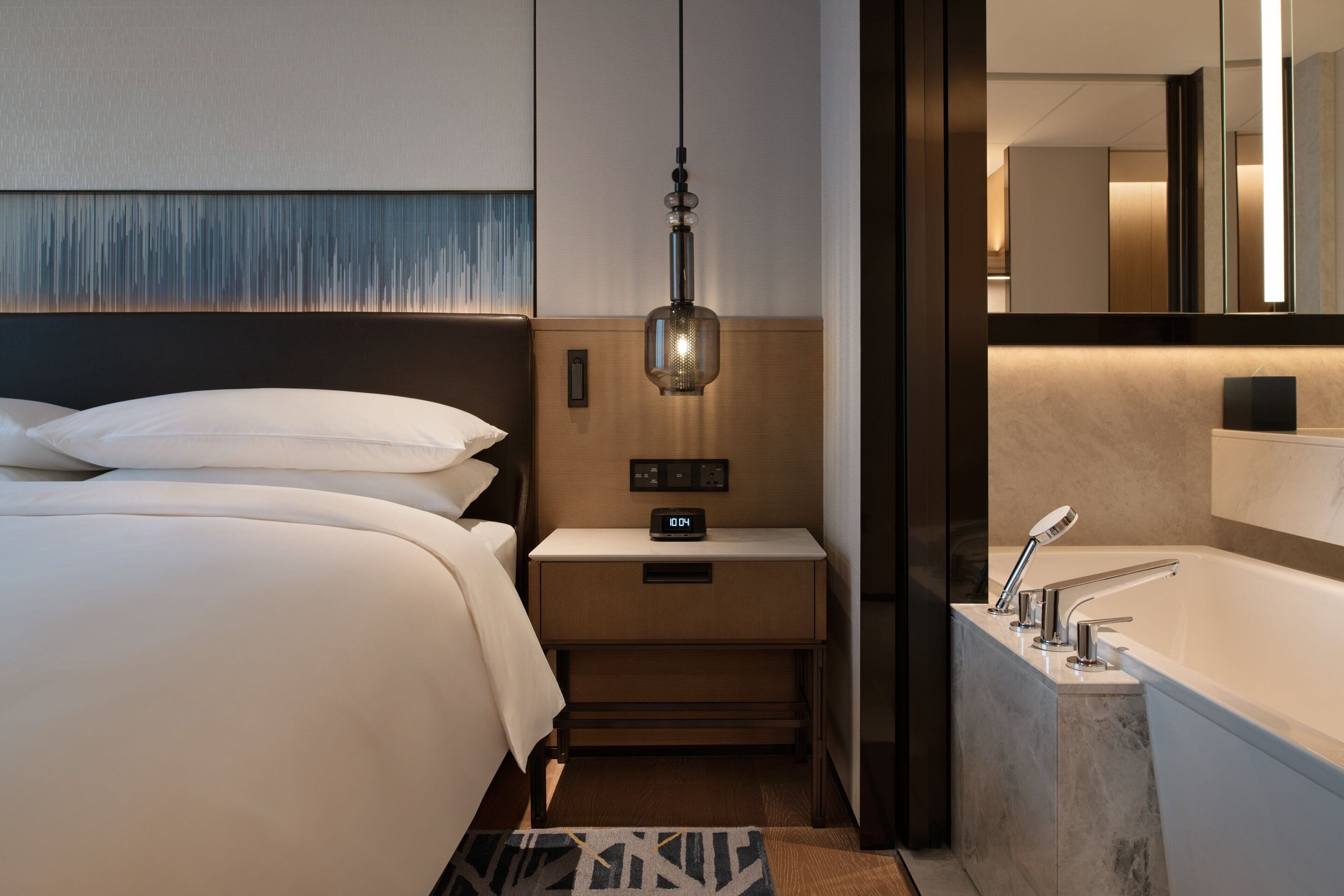 A modern hotel room with a large bed, bedside table with a clock, a bathroom with a bathtub, and a hanging light fixture.