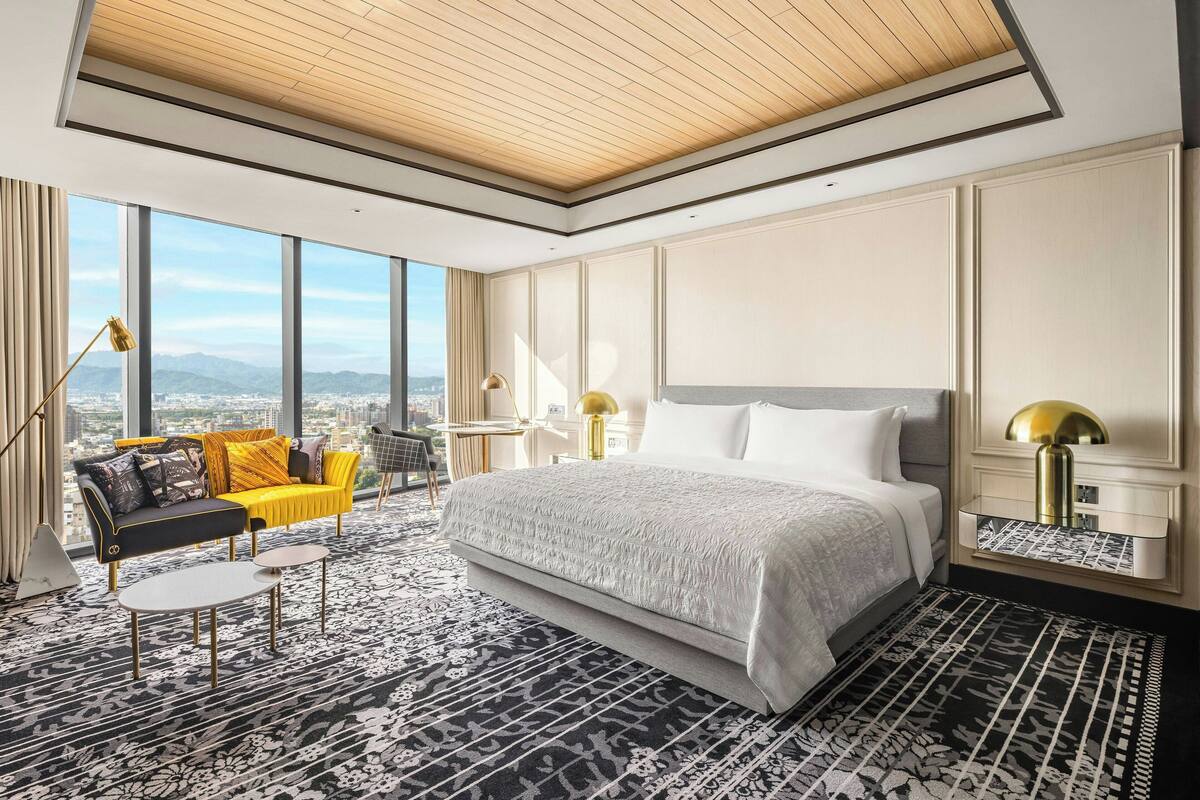 A modern hotel room with a large bed, a sofa, and a desk. The room has large windows offering a city view.
