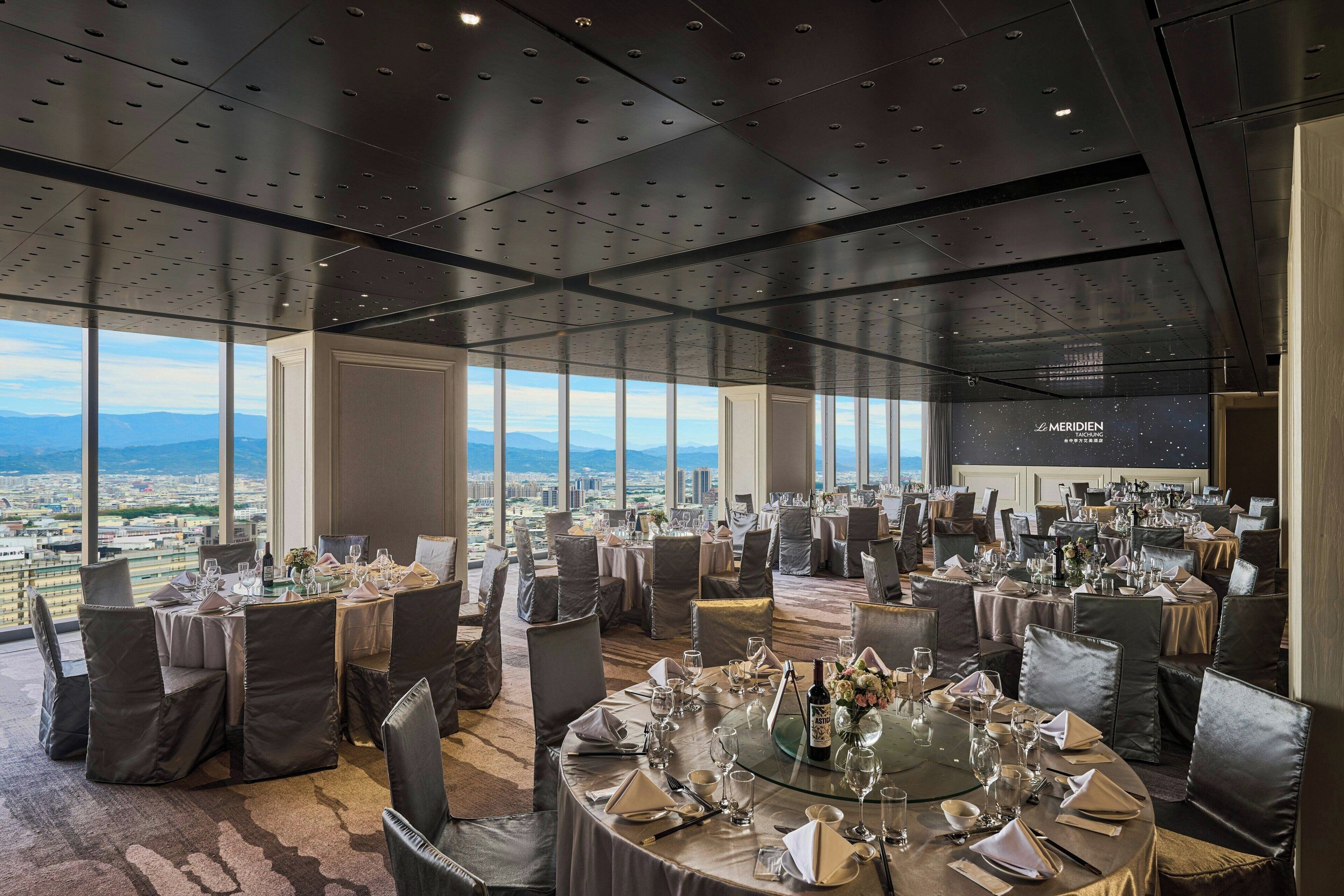 A high-rise dining area with round tables set for a meal, large windows offering a city view, and a modern ceiling design.