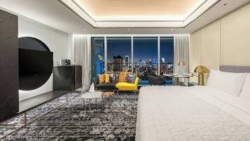 A modern hotel room with a large bed, a sofa, a coffee table, and a city view through large windows.