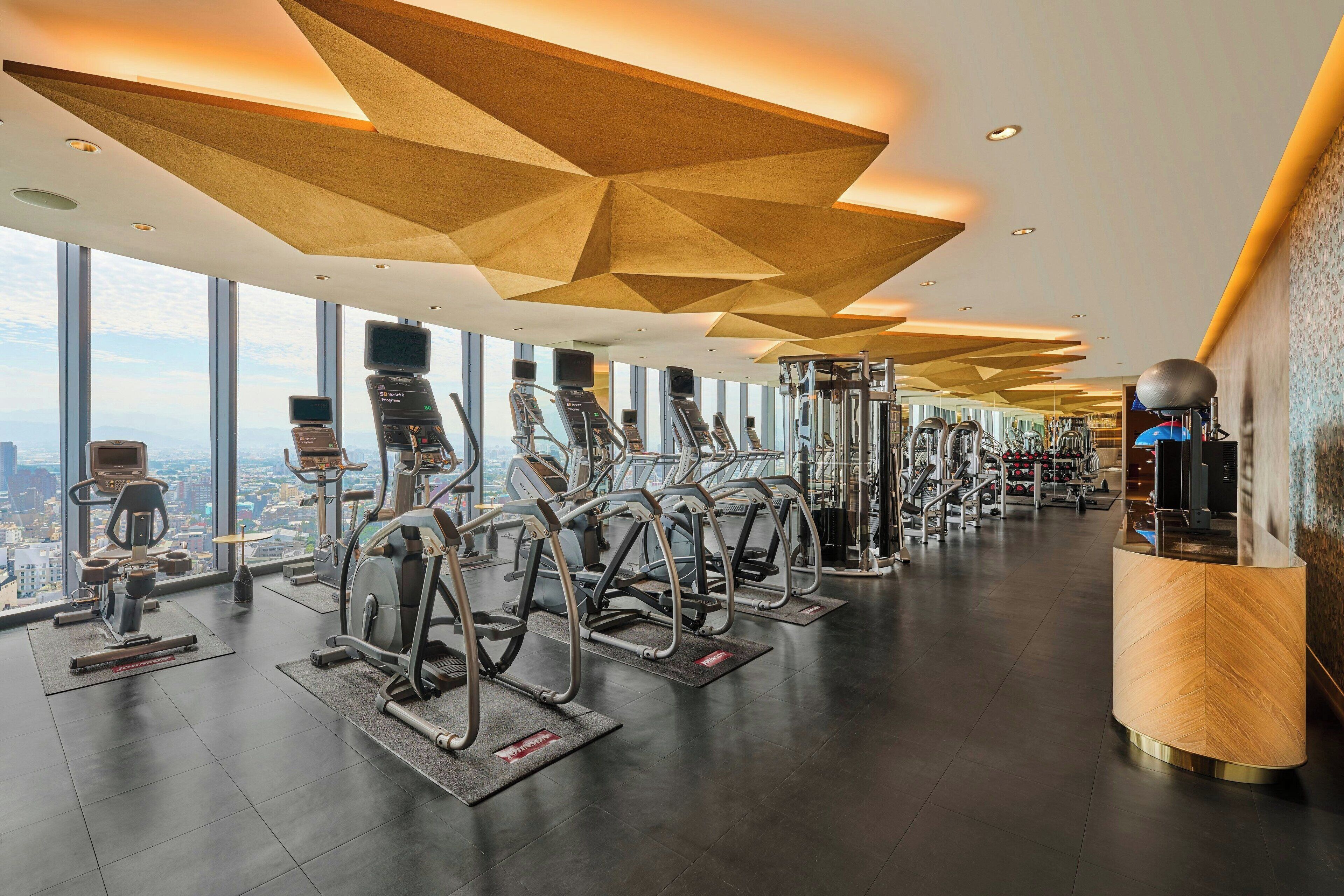 A modern gym with various exercise equipment, large windows offering city views, and a unique geometric ceiling design.