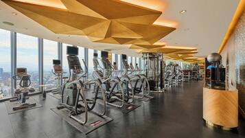 A modern gym with various exercise equipment, large windows offering city views, and a unique geometric ceiling design.