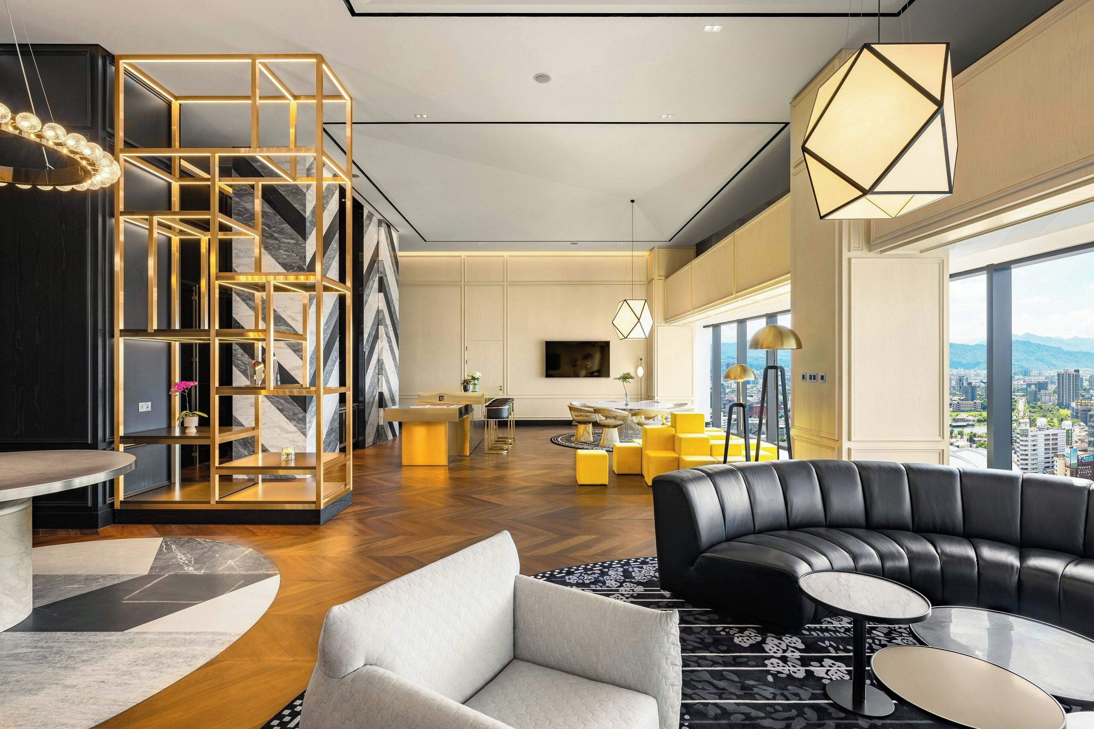 A modern living room with a black leather sofa, a white armchair, and a round coffee table. There is a geometric bookshelf and a large window offering a city view.