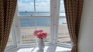 A room with a bay window overlooking a beach, featuring two white chairs and a vase of flowers.