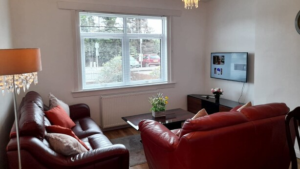 A living room with a red leather sofa, a small table with a vase, a floor lamp, a large window with blinds, and a TV on the wall.