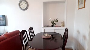 A dining area with a round table, four chairs, a clock on the wall, a red sofa, and a small shelf with decorative items.