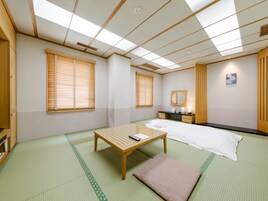 A room with tatami mats, a bed, a wooden table, a bedside table, a mirror, and a window with blinds.