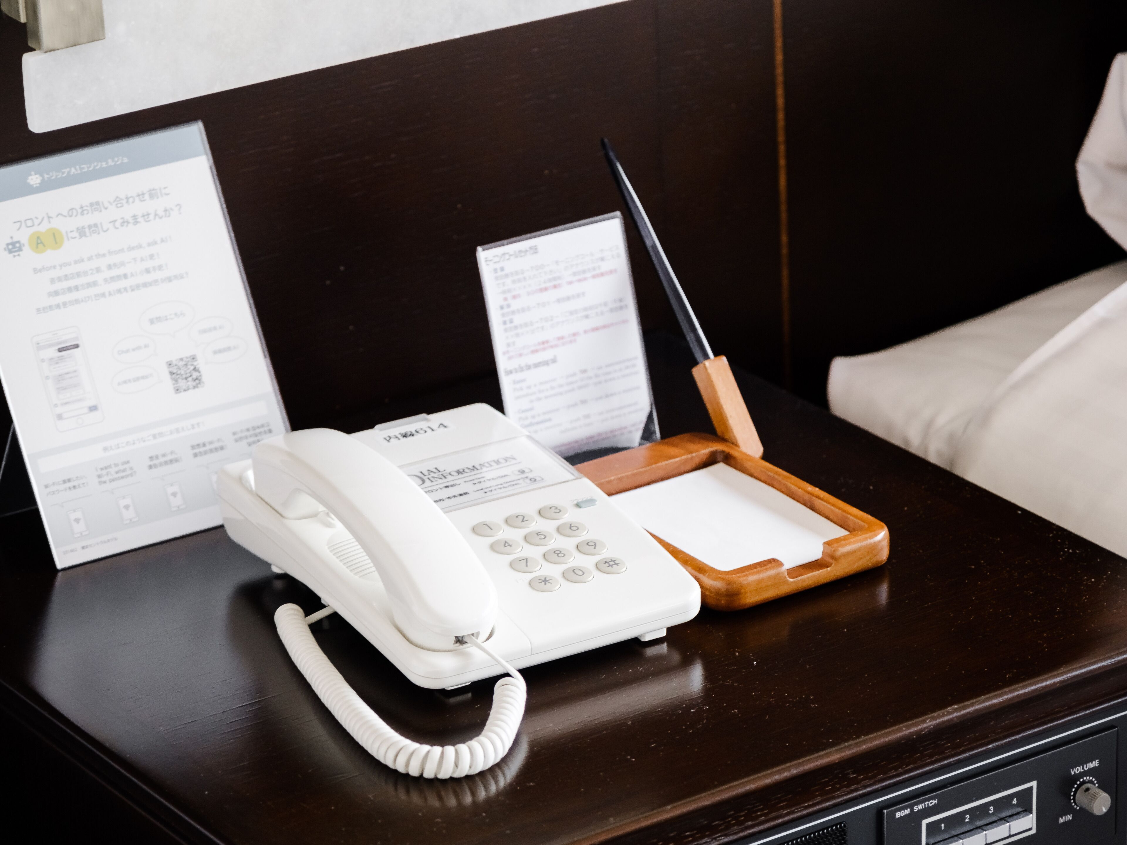 A hotel room with a wooden desk, a white telephone, a pen, and papers on the desk.
