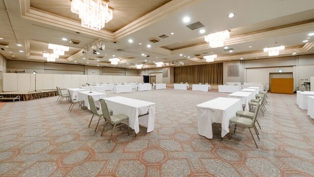 Uma ampla sala de conferências com várias mesas e cadeiras, um palco com um púlpito e um grande fundo de cortina.