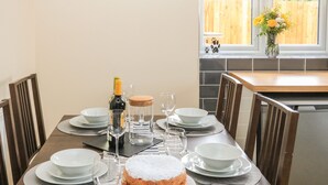 Ruokapöytä on katettu valkoisilla lautasilla, kulhoilla ja aterimilla. Pöydällä on myös tomusokerilla kuorrutettu kakku, viinipullo ja lasipurkki.