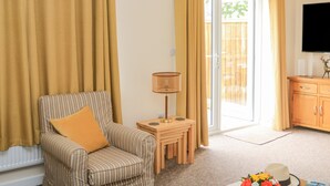 Uma sala de estar com uma poltrona listrada, uma pequena mesa de madeira com uma luminária, uma televisão sobre um móvel de madeira e uma porta de vidro deslizante que dá para uma área externa.
