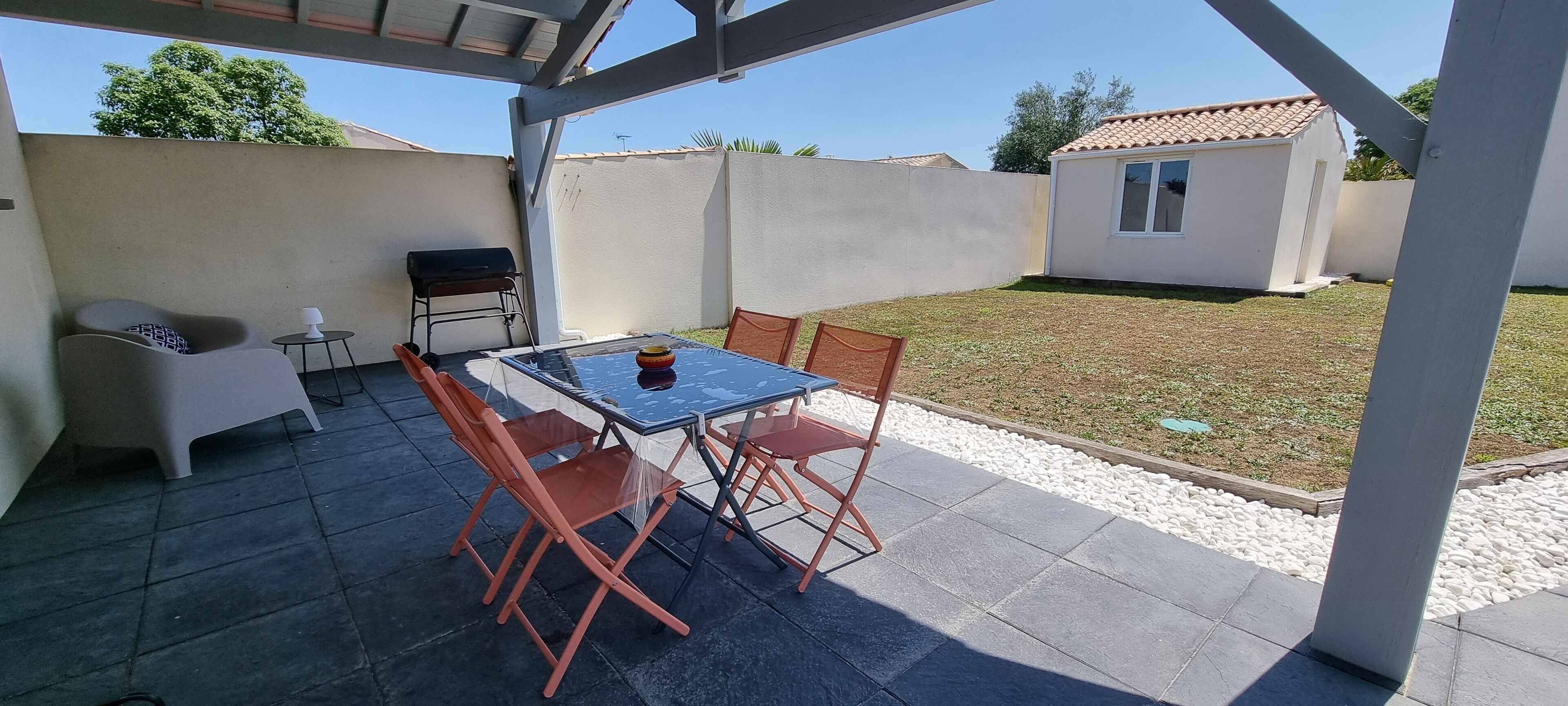 Ein überdachter Patio-Bereich mit Tisch und Stühlen, einem Grill und einem kleinen weißen Gebäude im Hintergrund.