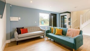 A living room with a grey sofa and a teal sofa, both with cushions. There is a window with a view of greenery outside. - Fully Accessible-disabled Adapted-hot Tub-pets (Alcester)