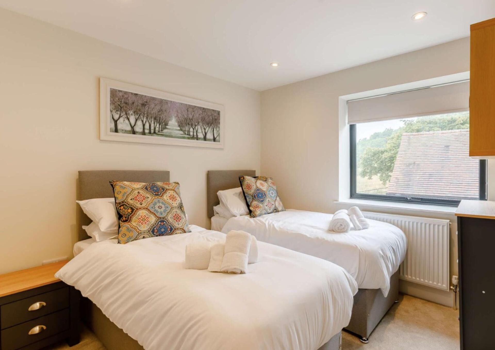 A bedroom with two beds, a window with a view, a radiator, and a painting on the wall.
