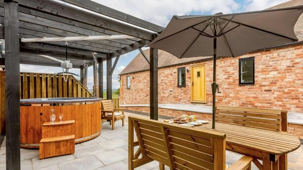 En udendørs terrasse med et træbord og bænke, et spabad samt en pergola med stor parasol.