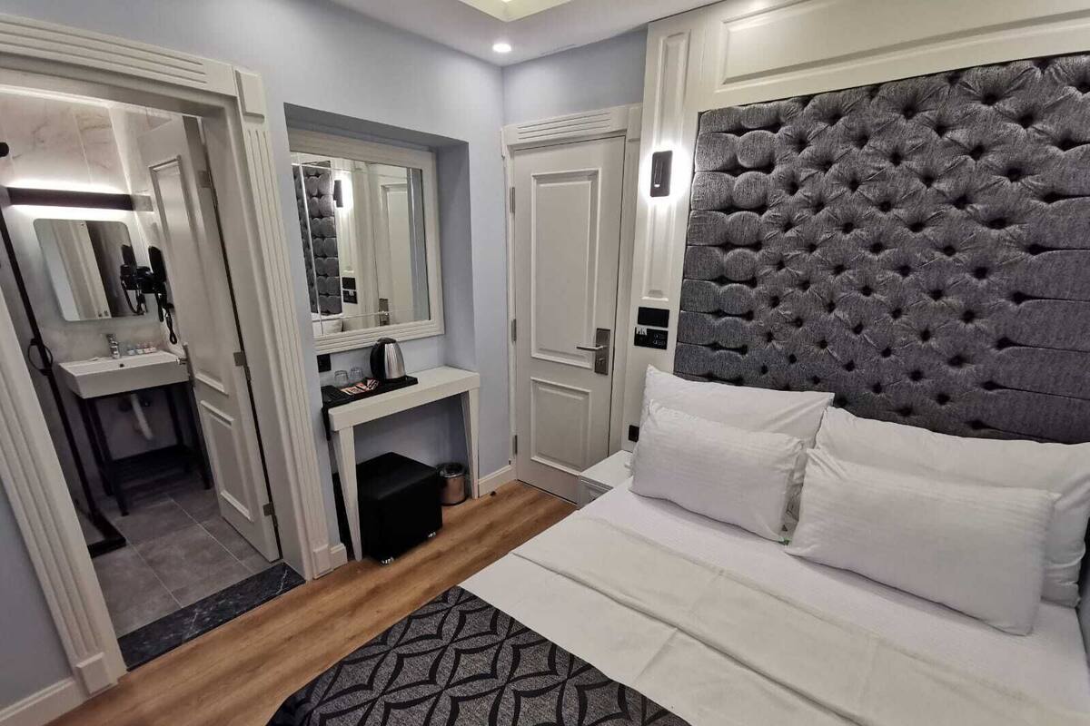 A bedroom with a bed, a bedside table, a mirror, a wardrobe, and a bathroom visible through an open door.