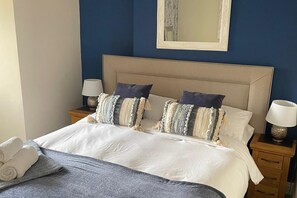 Un lit bien fait, avec du linge de lit blanc, deux oreillers et un plaid bleu. Il y a deux tables de chevet avec des lampes, et un miroir est accroché au mur au-dessus du lit.