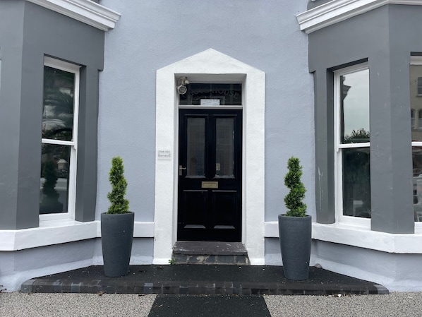 L’entrée d’un immeuble avec une porte noire, deux plantes en pot et une enseigne indiquant « CRIME RISE APARTMENTS ».