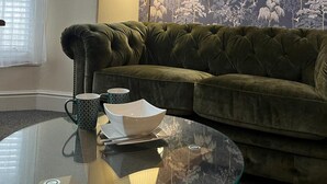Un salon avec un canapé en velours vert, une table basse en verre sur laquelle reposent un bol blanc et deux tasses à motifs, et un papier peint à motifs floraux.