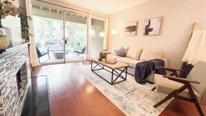 A modern living room with a wooden floor, a sofa, a coffee table, and a fireplace. There is a sliding glass door leading to an outdoor area with a chair and a table.