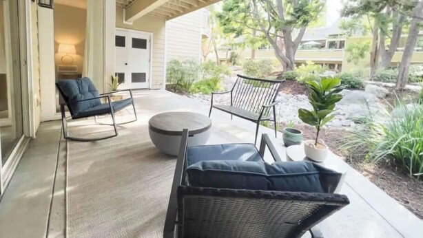 A covered patio with two black wicker chairs and a round table, a door leading inside, and a garden with trees and plants.