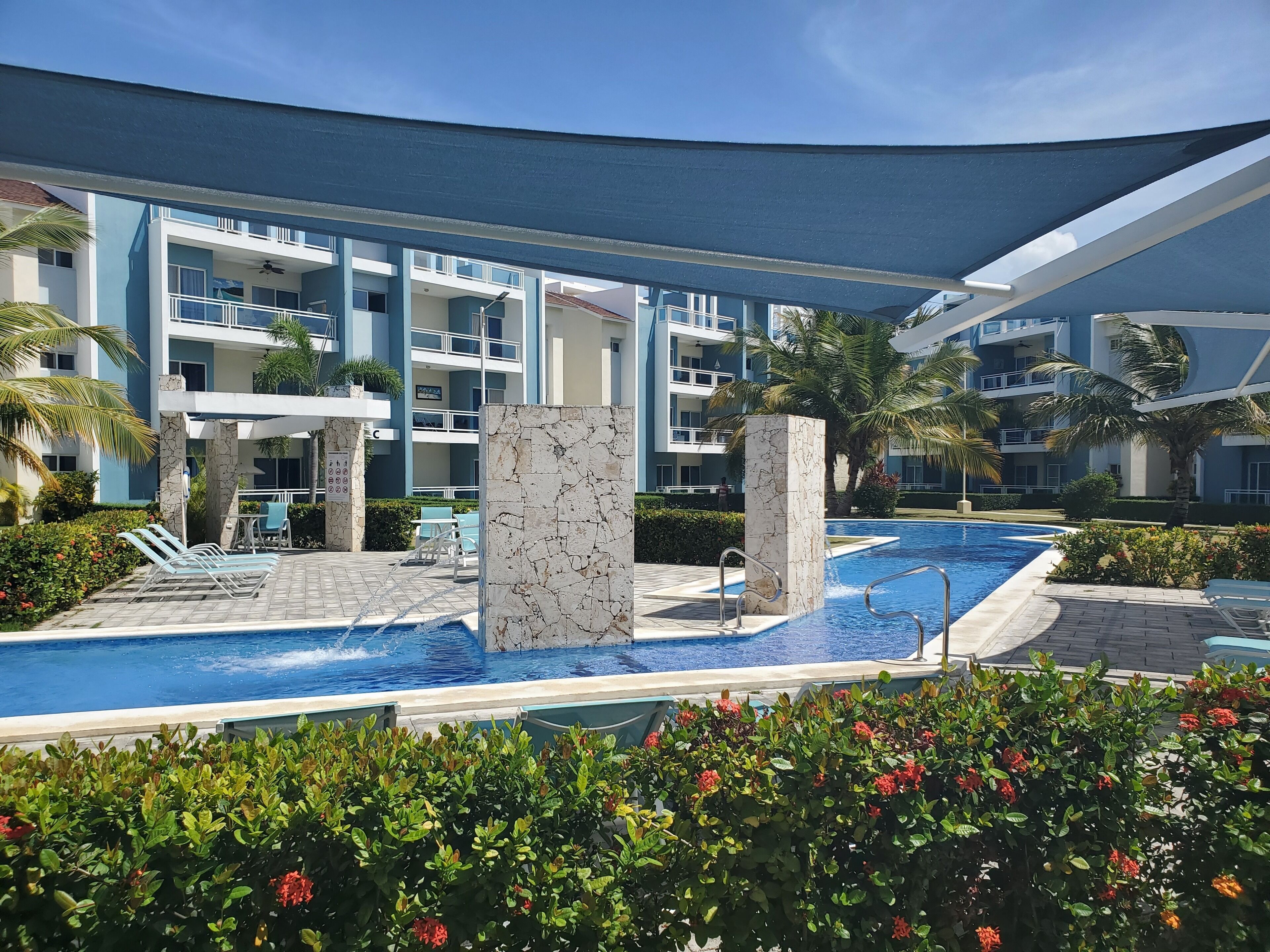Area piscina di un hotel, con una piscina azzurra e cristallina, lettini e una palma.