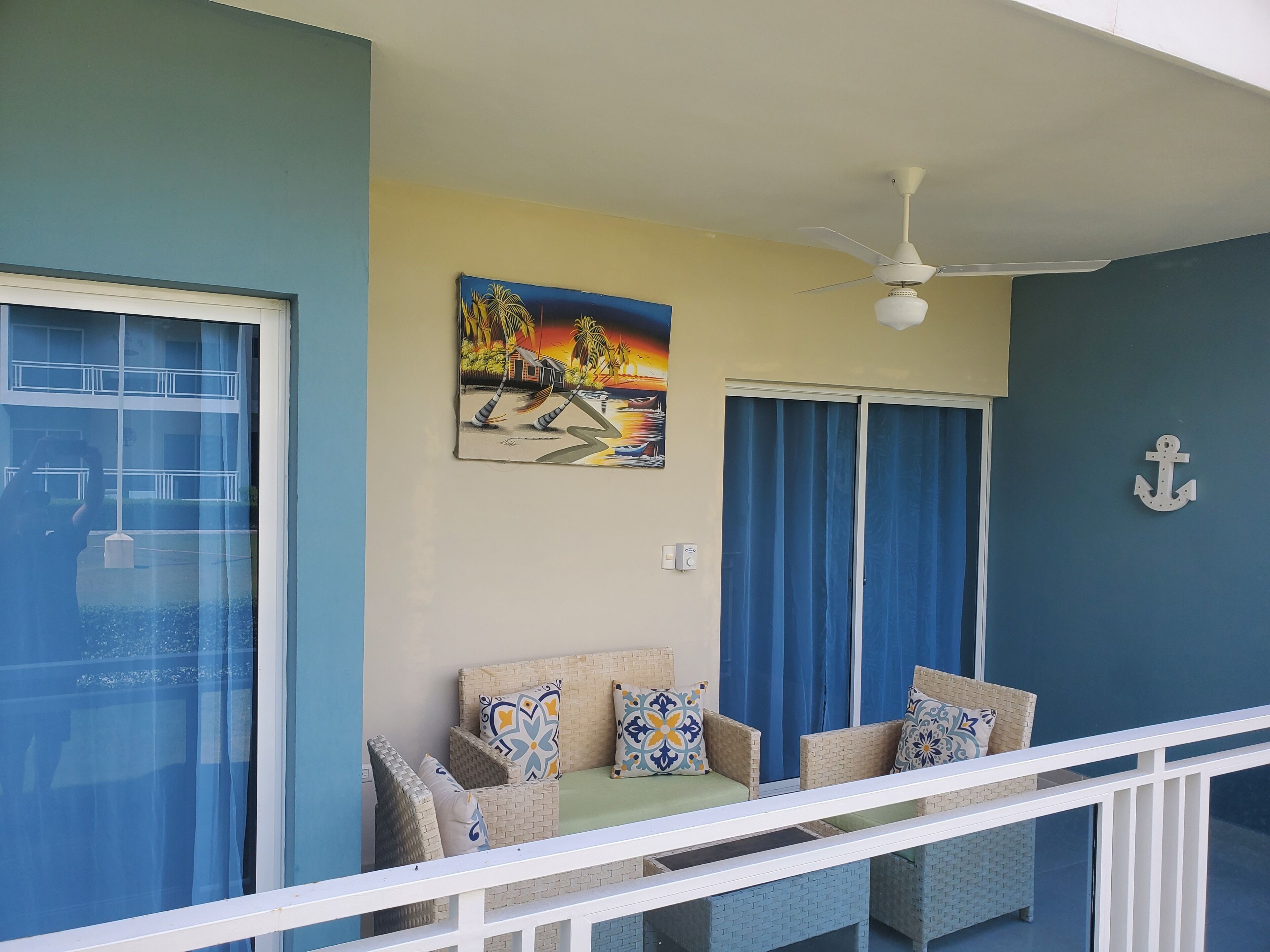 A balcony with wicker furniture, a ceiling fan, and a painting of a sunset scene.